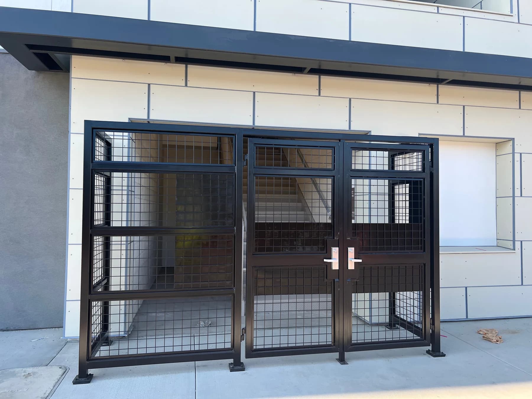 Black welded-mesh security enclosure with paired access doors beside a commercial building