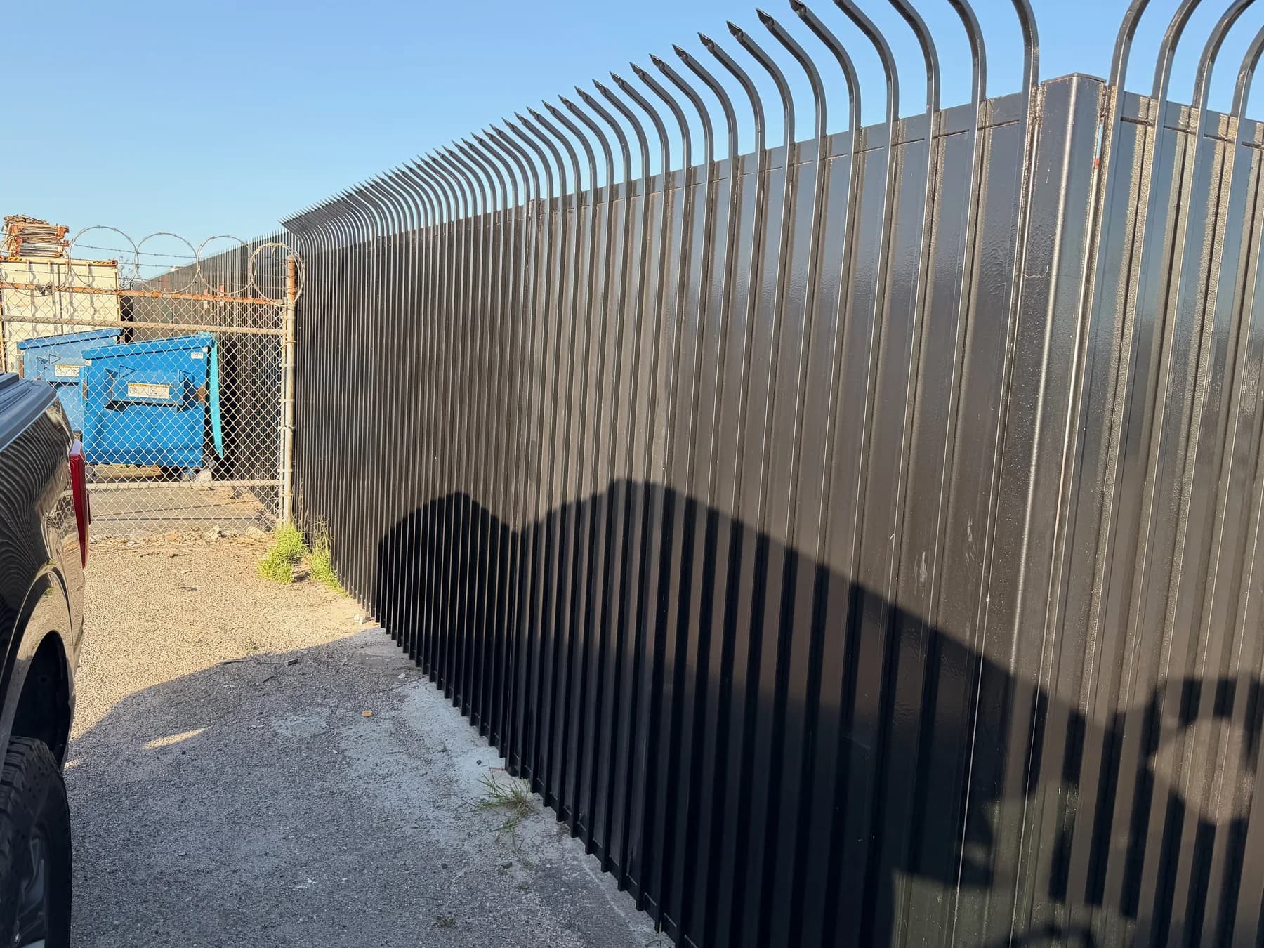 Tall black privacy fence with outward-curved anti-climb pickets along an industrial yard