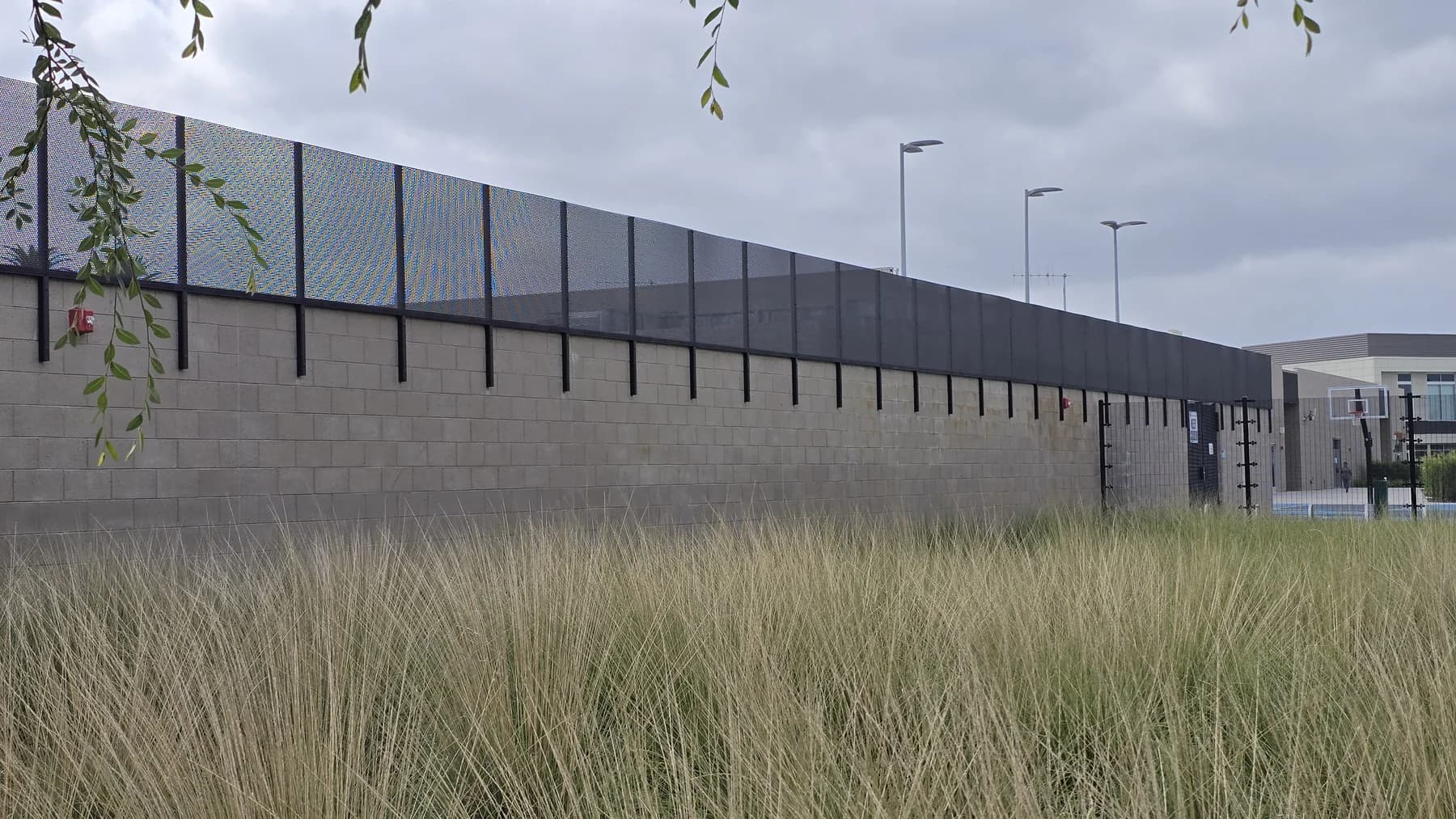 Welded-mesh security extension above a long masonry wall beside landscaped grounds
