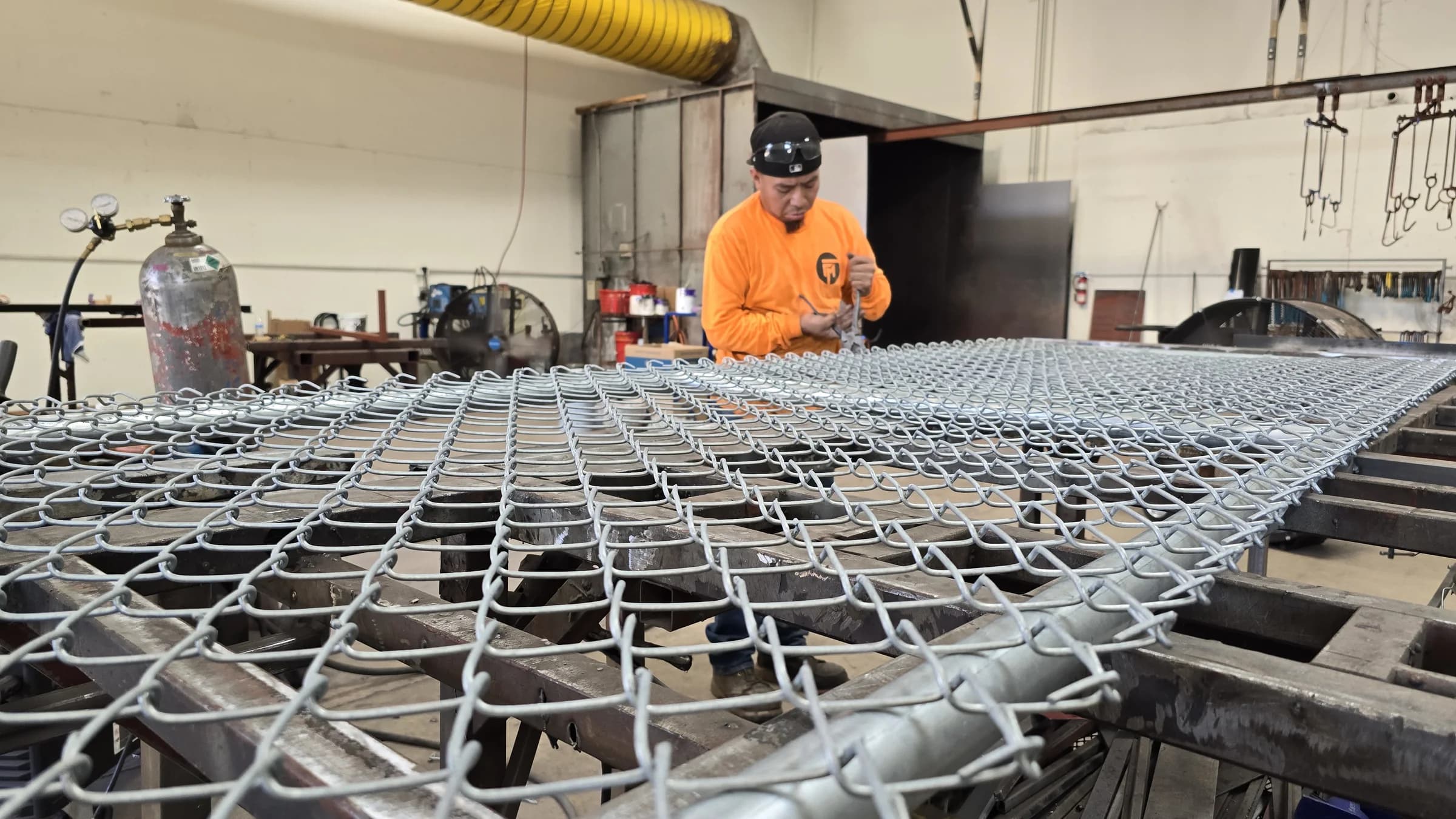 Fence Menders fabricator with gloved hands securing chain-link mesh to a gate frame